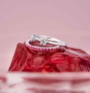 Zwei silberne Ringe liegen auf einer roten, facettierten Oberfläche. Ein Ring ist mit einem klaren Edelstein versehen, während der andere mit kleinen roten Steinen geschmückt ist. Der Hintergrund ist in zartem Rosa gehalten und schafft eine romantische und elegante Atmosphäre – perfekt für jede Hochzeitsfeier oder Heiratsinspiration.