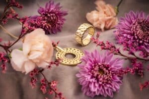 Ein goldener Ring und ein Anhänger mit komplizierten Mustern werden zwischen rosa Nelken, lila Chrysanthemen und zarten roten Blumen auf einer sanften grauen Oberfläche präsentiert, perfekt für alle, die gerade ihre Hochzeit planen.