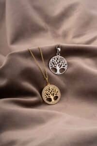 Zwei runde Anhänger mit Baummotiven ruhen auf einem weichen, beigen Stoff. Perfekt für eine Hochzeit: Ein Anhänger ist golden mit einer passenden Kette, der andere ist silbern mit einer passenden Kette, beide mit aufwendigen Baumausschnitten. Ideale Andenken für Ihren besonderen Hochzeitstag.