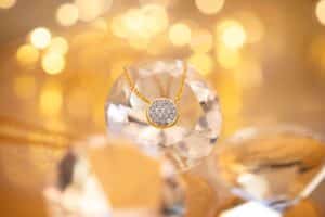Eine goldene Halskette mit einem runden, diamantbesetzten Anhänger wird elegant auf einem großen, facettierten Kristall präsentiert. Der Hintergrund, der an Hochzeitsplanungsträume erinnert, ist mit verschwommenen, warmen goldenen Lichtern geschmückt, die eine luxuriöse und festliche Atmosphäre schaffen.