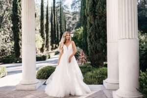 Eine Frau in einem fließenden weißen Brautkleid steht zwischen großen Säulen, umgeben von üppigem Grün und hohen Bäumen. Während sie von ihrer perfekten Hochzeit träumt, blickt sie zur Seite und schafft so eine heitere und elegante Szene in dieser malerischen Außenkulisse.