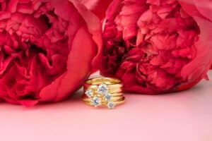 Ein Stapel goldener Ringe mit Diamanten schimmert vor zwei großen, leuchtend rosa Pfingstrosen auf einem pastellrosa Hintergrund und fängt die Essenz einer Hochzeit ein.