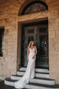Eine Frau in einem fließenden weißen Hochzeitskleid steht auf den Stufen eines großen Steingebäudes mit kunstvollen Metalltüren und Bogenfenstern und fängt die Essenz einer perfekten Hochzeit ein.