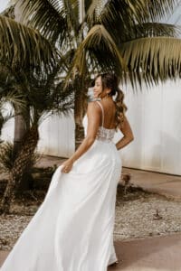 Eine Frau in einem weißen Brautkleid geht davon und hält dabei den Rock ihres Kleides. Sie hat langes, welliges Haar und ein strahlendes Lächeln. Palmen rahmen die Szene als Teil dieser magischen Hochzeit ein, und ein helles Gebäude verleiht diesem bildschönen Moment des Heiratens Charme.