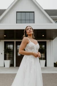 Eine Frau in einem weißen Hochzeitskleid steht vor einem modernen Gebäude mit großen Fenstern. Sie blickt leicht nach oben, ihr langes Haar umrahmt ihr Gesicht und sie faltet die Hände. Das Kleid, perfekt für jede Hochzeit, weist atemberaubende florale Spitzendetails auf.