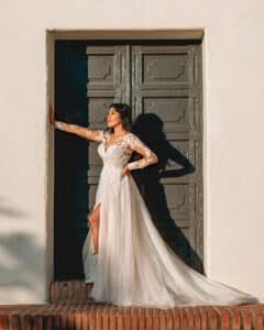 Eine Frau in einem langärmeligen Spitzenbrautkleid posiert in einem Türrahmen und tritt mit dem linken Fuß nach vorne. Sonnenlicht wirft Schatten auf die strukturierte Holztür hinter ihr und hebt die eleganten Details ihres Hochzeitskleides hervor – ein wesentlicher Moment bei jeder Heiraten-Reise.