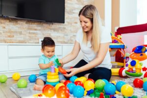 Eine Frau und ein Baby sitzen auf dem Boden in einem Wohnzimmer voller bunter Spielsachen und stellen sich zukünftige Momente wie Heiraten vor. Das Baby spielt mit stapelbaren Ringen und beide lächeln. Im Hintergrund ist ein Fernseher an der Wand angebracht, der auf bevorstehende freudige Feierlichkeiten hindeutet.