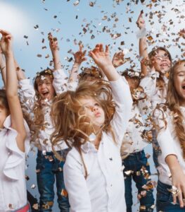 Eine Gruppe fröhlicher Kinder in weißen Hemden und Jeans wirft vor einem hellblauen Hintergrund Konfetti in die Luft. Sie lächeln und feiern eine Hochzeit, während sie von Konfetti umgeben sind, während sie springen und lachen.