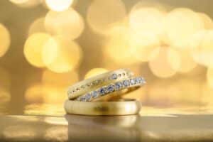 Zwei Goldringe sind kunstvoll vor einem verschwommenen goldenen Hintergrund gestapelt und fangen die Essenz einer Hochzeit ein. Ein Ring hat eine glatte Oberfläche, während der andere mit einer Reihe funkelnder Diamanten verziert ist. Die Szene strahlt Eleganz und Luxus aus, perfekt für Hochzeitsträume.