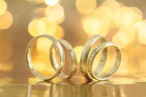 Ein Set eleganter Goldringe mit unterschiedlichen Designs, darunter glatte und strukturierte Oberflächen, einige mit kleinen Diamanten verziert. Perfekt für die Hochzeitsplanung, werden sie vor einem warmen, verschwommenen Hintergrund mit kreisförmigem Licht-Bokeh präsentiert und schaffen so eine luxuriöse und romantische Atmosphäre.
