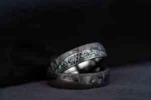 Nahaufnahme von zwei eleganten schwarzen Ringen auf einer dunklen Oberfläche. Der obere Ring ist perfekt für Ihre Hochzeit und verfügt über ein Band aus kleinen, funkelnden blauen und weißen Steinen, die ihm einen Hauch von Eleganz verleihen. Der sanft verschwommene Hintergrund hebt die komplizierten Details der Ringe hervor.