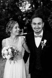 Ein Schwarzweißfoto fängt die Essenz einer Hochzeit ein: Die Braut lacht, den Blumenstrauß in der Hand, und trägt ein ärmelloses Kleid. Der Bräutigam, Anzug und Fliege tragend, lächelt herzlich, während sie Hand in Hand zwischen den Bäumen spazieren – perfekte Momente, die durch eine durchdachte Hochzeitsplanung entstehen.