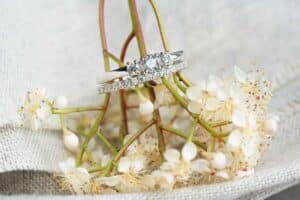 Zwei funkelnde Ringe, einer mit einer Reihe Diamanten und der andere mit einem einzelnen Diamanten, ruhen auf zarten weißen Blumen. Der Hintergrund ist ein weicher, strukturierter Stoff, der das elegante und romantische Ambiente verstärkt – perfekt für Ihre Hochzeits- oder Hochzeitsplanungsträume.