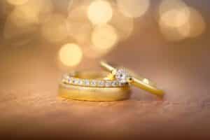 Eine Nahaufnahme von zwei Goldringen auf einer weichen, strukturierten Oberfläche fängt die Essenz der Hochzeitsplanung ein. Ein Ring ist mit einer Reihe funkelnder Diamanten verziert, während der andere mit einem einzelnen runden Diamanten geschmückt ist. Der Hintergrund ist sanft verschwommen mit warmen, leuchtenden Bokeh-Lichtern, perfekt für Ihre Hochzeitsträume.