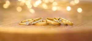Eine Sammlung goldener Ringe, einige davon mit Diamanten verziert, sind elegant auf einer Holzoberfläche arrangiert. Weiches, verschwommenes Licht schafft einen warmen, leuchtenden Hintergrund, perfekt für Inspirationen bei der Hochzeitsplanung.