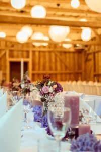 Eine rustikale Hochzeitstafel mit Arrangements aus lila und rosa Blumen, Kerzen und Weingläsern. Der Hintergrund besteht aus einem Holzinterieur mit sanften, leuchtenden Hängelampen – perfekt für alle, die gerade mitten in der Hochzeitsplanung stecken.