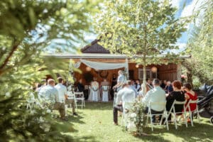 Eine Hochzeitszeremonie im Freien findet statt, bei der die Gäste auf weißen Stühlen mit Blick auf den Altar sitzen. Das Paar steht unter einer hölzernen Pergola, die mit weißen Vorhängen geschmückt ist. Umgeben von Grün vervollständigt ein rustikales Gebäude diese idyllische Kulisse, perfekt für eine durchdachte Hochzeitsplanung.