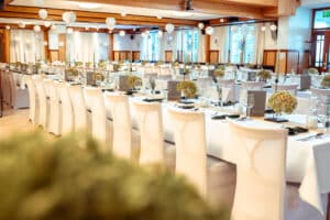 Ein großer Bankettsaal mit langen Tischen, die für eine Hochzeit gedeckt sind. Jeder Tisch ist mit kleinen Blumenarrangements und eleganten Gedecken dekoriert. Weiße Stühle und sanfte Beleuchtung schaffen eine einladende Atmosphäre, die perfekt für die Hochzeitsplanung ist, während Papierlaternen von der Decke hängen.