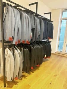An einem Kleiderständer im Bekleidungsgeschäft werden elegante Herrenanzüge in Grau-, Schwarz- und Blautönen ausgestellt, perfekt für jede Hochzeit. Sie hängen ordentlich an Metallstangen mit roten Preisschildern. Der Laden verfügt über Holzboden und ein großes Fenster und ist somit ein idealer Anlaufpunkt für alle, die gerade mitten in der Hochzeitsplanung stecken.