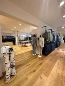 Ein gut beleuchteter Bekleidungsladen mit Holzboden, perfekt für alle, die gerade ihre Hochzeit planen. Ordentlich angeordnete Anzüge zieren die Kleiderständer und Schaufensterpuppen und bieten eine elegante Auswahl für alle, die heiraten möchten. Der geräumige, organisierte Bereich präsentiert zusätzliche Anzüge auf Kleiderständern im Hintergrund.