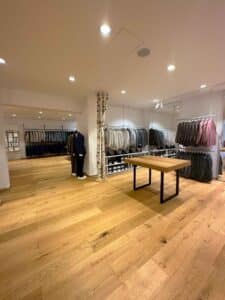Ein modernes Bekleidungsgeschäft mit Holzfußboden bietet eine Auswahl an Anzügen und Hemden in neutralen Farbtönen, perfekt für die Hochzeitsplanung. In der Mitte steht ein Holztisch, ideal zum Durchstöbern von Hochzeitskleidern, da der Raum mit Deckenleuchten gut beleuchtet ist, um jedes Detail hervorzuheben.