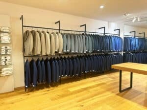 Im Inneren eines Bekleidungsgeschäfts werden ordentlich arrangierte Anzüge in Beige-, Grau- und Blautönen präsentiert – perfekt für jede Hochzeit. Im Vordergrund steht ein Holztisch, auf den Regalen links davon liegen gefaltete Hemden. Der helle Holzboden trägt zur eleganten Atmosphäre bei, die ideal für die Hochzeitsplanung ist.