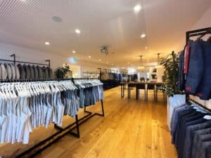 Ein modernes Bekleidungsgeschäft mit ordentlich angeordneten Kleiderständern voller Kleidung, darunter Tanktops und Jacken für Ihre Flitterwochen. Der Laden hat Holzboden, helle Beleuchtung und ein minimalistisches Design. In der Mitte steht ein langer Tisch mit Hockern, der das Set-up ideal für Diskussionen nach der Hochzeitsplanung macht.