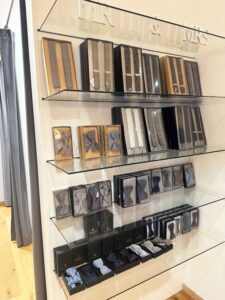 Ein Ausstellungsregal präsentiert eine Auswahl verpackter Krawatten und Fliegen in verschiedenen Farben und Mustern, perfekt für Hochzeitsveranstaltungen. Ordentlich in Schachteln auf Glasregalen vor einer hellen Wand angeordnet, sind sie ideal für jede Hochzeitsplanung. Der Holzboden und ein Teil eines Vorhangs sind ebenfalls sichtbar.