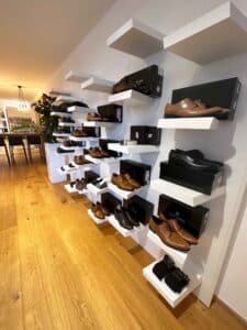 Ein modernes Schuhgeschäft mit weißen Wandregalen zeigt eine Auswahl an Herrenschuhen in Braun und Schwarz, perfekt für die Hochzeitsplanung. Die Schuhe sind ordentlich in ihren Kartons auf dem Holzboden angeordnet, während im Hintergrund ein langer Tisch sichtbar ist.