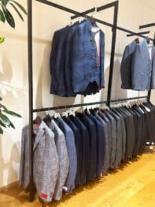 Auf einem Kleiderständer hängen mehrere blaue und graue Herrenblazer, perfekt für die Hochzeitsplanung. In der oberen Reihe hängen Blazer mit gemusterten Designs, während in der unteren Reihe ordentlich unifarbene und gemusterte Blazer zu sehen sind. Die Kulisse ist ein Laden mit elegantem Holzboden.