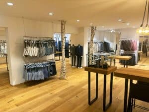 Ein Bekleidungsgeschäft mit ordentlich angeordneten Herrenhemden und Anzügen auf Ständern, perfekt für alle, die sich auf Hochzeitsplanung konzentrieren. Der Holzboden verleiht dem modernen, gut beleuchteten Interieur Wärme. Tische im Vordergrund stellen Accessoires zur Schau, während Hängelampen eine gemütliche Atmosphäre schaffen.