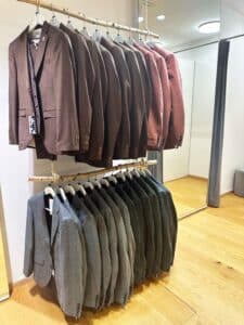 In einem Bekleidungsgeschäft sind zwei Kleiderständer mit Herrenanzügen zu sehen, die sich ideal für die Hochzeitsplanung eignen. Der obere Ständer enthält braune Anzüge, der untere graue Anzüge. Die Anzüge sind ordentlich auf Kleiderbügeln an einer weißen Wand mit Holzboden darunter aufgereiht – die perfekte Kleidung für diejenigen, die heiraten möchten.
