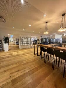 Ein modernes Ladeninterieur mit Holzboden, Kleiderständern und Regalen, in denen gefaltete weiße Hemden ausgestellt sind. Schwarze Barhocker sind um einen Holztisch herum angeordnet. Perfekt, um Hochzeitskleidung auszuprobieren, während warme Hängelampen eine gemütliche Atmosphäre schaffen, ideal für jede Hochzeitsplanungssitzung.