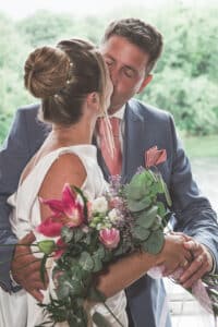 Bei ihrer Hochzeit küsst sich das Paar zärtlich. Die Braut hält einen Strauß rosa Lilien und üppiges Grün, während der Bräutigam in einem blauen Anzug mit rosa Krawatte schick aussieht. Sie stehen vor einem Fenster mit einer grünen, blattreichen Kulisse.