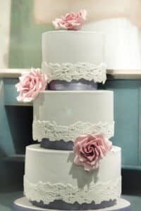 Eine dreistöckige weiße Hochzeitstorte, perfekt für Ihre Hochzeit, geschmückt mit zarten rosa Rosen und aufwendigen Spitzenmustern zwischen den einzelnen Schichten, präsentiert vor einem weichen, eleganten Hintergrund.