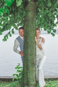 Ein Paar in formeller Kleidung schaut lächelnd hinter einem Baum hervor. Sie stehen an einem ruhigen See, über dem sich grüne Blätter befinden. Der Mann trägt einen Anzug und die Frau ein weißes Kleid – ein idyllischer Moment, perfekt für die Hochzeitsplanung vor der ruhigen Wasserlandschaft.