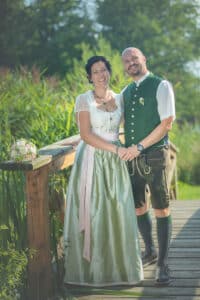 Ein Paar steht auf einer Holzbrücke, umgeben von üppigem Grün, und strahlt, während sie Händchen halten. Die Frau trägt ein traditionelles Kleid mit einem grünen Rock, während der Mann eine grüne Weste über einem weißen Hemd und Lederhosen trägt. Es ist ein bildschöner Moment für ihre Hochzeit, und Details der Hochzeitsplanung wie der Blumenstrauß verleihen dem Ganzen Charme.