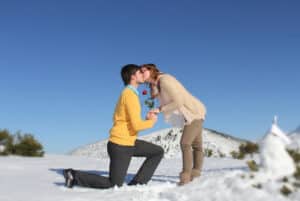 Eine Person kniet im Schnee und bietet einer anderen stehenden Person eine Rose an, während sie sich küssen – ein malerischer Moment, umgeben von schneebedeckten Hügeln und einem klaren blauen Himmel. Die Szene wirkt wie ein bezaubernder Auftakt ihrer Hochzeitsplanung, und beide sind für diesen Anlass warm gekleidet.