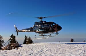 Ein schwarzer Hubschrauber schwebt über dem schneebedeckten Gelände und erinnert an einen aufregenden Auftritt bei einer Winterhochzeit, mit klarem blauen Himmel im Hintergrund. Hohe Kiefern stehen majestätisch auf dem schneebedeckten Boden.