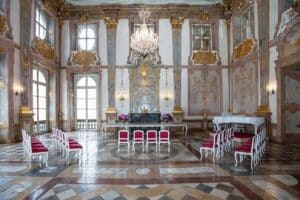Ein großer, kunstvoller Ballsaal mit hohen Decken, großen Fenstern und einem Kronleuchter bildet die Bühne für eine unvergessliche Hochzeit. Der Raum verfügt über dekorative Goldleisten und einen Marmorboden. Weiße Stühle mit roten Kissen sind in Halbkreisen angeordnet und suggerieren eine formelle Veranstaltungsanordnung, die perfekt für die Hochzeitsplanung ist.