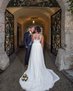 Braut und Bräutigam stehen in einem gewölbten Gang mit verzierten Toren und fangen damit perfekt den Geist einer Hochzeit ein. Die Braut in einem weißen Kleid hält einen Blumenstrauß und blickt abgewandt, während der Bräutigam in einem dunklen Anzug sich lächelnd zu ihr beugt. Über ihnen hängt ein Laternenlicht.