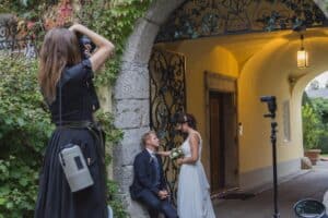 Ein Fotograf fängt einen Hochzeitsmoment ein, in dem Braut und Bräutigam unter einem dekorativen Torbogen posieren. Die Braut in einem weißen Kleid hält einen Blumenstrauß, während der Bräutigam auf einem Sims sitzt. Üppiges Grün umgibt sie und eine Laterne hängt vom Bogen herab und bildet den perfekten Rahmen für diese romantische Hochzeitsszene.
