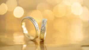 Zwei silberne Eheringe mit strukturierter Oberfläche werden auf einer reflektierenden goldenen Oberfläche präsentiert. Der Hintergrund zeigt warmes, verschwommenes goldenes Licht und fängt die Essenz einer romantischen und eleganten Hochzeitsatmosphäre ein.