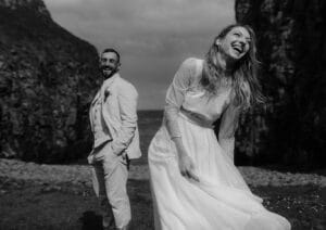 Ein glückliches Paar posiert im Freien zwischen felsigen Klippen und feiert seine Hochzeit. Die Frau, gekleidet in ein langes weißes Kleid, lacht und beugt sich leicht nach vorne, während der Mann im Anzug lächelnd hinter ihr steht. Die Szene ist in zeitlosem Schwarzweiß festgehalten.
