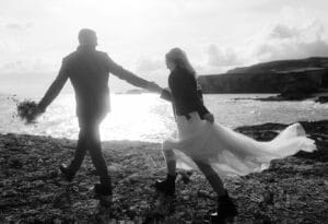 Ein Schwarzweißfoto fängt die Essenz einer Hochzeit ein, bei der Braut und Bräutigam Hand in Hand an einer felsigen Küste entlanggehen. Die Braut hält vorsichtig ihr Kleid, während der Bräutigam einen Blumenstrauß trägt. Das Meer und die Klippen bilden eine atemberaubende Kulisse für ihren besonderen Tag.