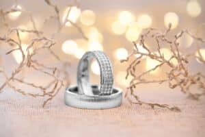 Zwei silberne Eheringe ruhen auf einer strukturierten Oberfläche und verkörpern die Essenz einer Hochzeit. Ein Ring ist schlicht, während der andere mit kleinen Diamanten verziert ist. Zarte Zweige rahmen die Ringe ein und weiche, unscharfe Lichter im Hintergrund schaffen eine warme, festliche Atmosphäre, die perfekt für die Hochzeitsplanung ist.