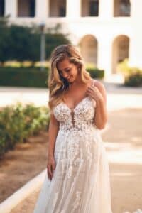 Eine Frau in einem Spitzenbrautkleid steht im Freien und verkörpert die Eleganz einer perfekten Hochzeitsplanung. Das Kleid weist florale Details und einen tiefen Ausschnitt auf. Sie hat langes, welliges blondes Haar und blickt mit einem sanften Lächeln nach unten. Der Hintergrund ist mit Grün und einem sonnenbeschienenen Gebäude verschwommen und verkörpert glückselig heiraten.