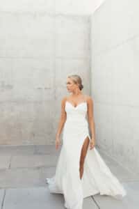 Eine Frau in einem weißen Hochzeitskleid steht in einer minimalistischen Betonkulisse und verkörpert die Einfachheit moderner Hochzeitsplanung. Das Kleid hat Spaghettiträger und einen hohen Schlitz. Sie hat kurzes blondes Haar und blickt zur Seite, was eine elegante und heitere Pose ergibt, die perfekt für Heiratsmomente ist.