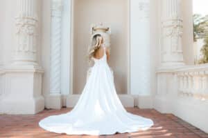 Eine Frau in einem fließenden weißen Kleid steht mit dem Rücken zur Kamera und verkörpert die Essenz von Hochzeit vor einer kunstvollen architektonischen Kulisse mit hohen Säulen und aufwendigen Details. Ihr langes blondes Haar fällt anmutig über ihren Rücken.