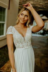 Eine Frau in einem weißen Spitzenkleid steht unter einem Torbogen und berührt sanft mit der Hand ihren Kopf. Sie lächelt sanft und verkörpert die Freude einer Hochzeit, wobei ihr blondes Haar locker auf ihre Schultern fällt. Im Hintergrund ist ein Gebäude mit einem Fenster und einem Steinweg zu sehen, das die Szene für die Hochzeitsplanung darstellt.